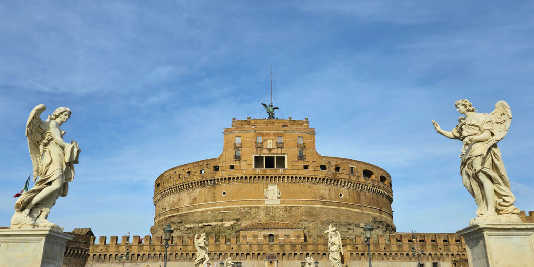 A vantage view of the cylindrical fortress flanked by two marble angel statues against a clear blue sky.