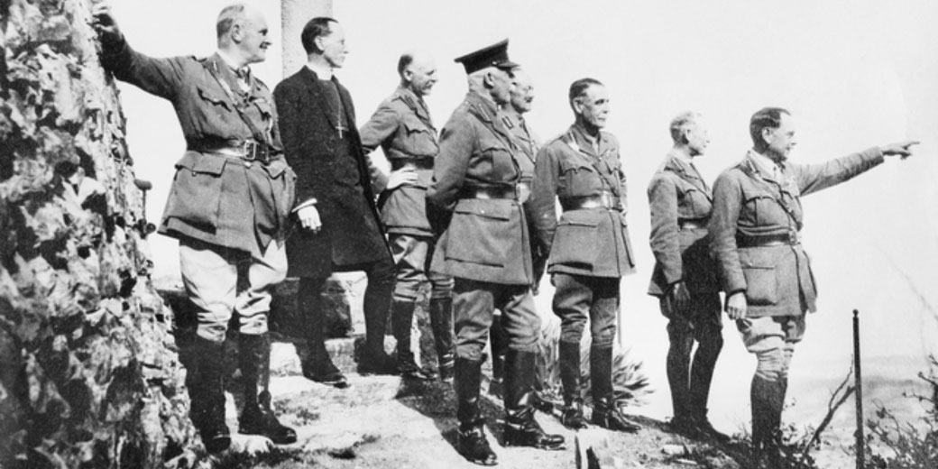 A group of military officers and a clergyman stand on a rocky ledge, with one officer at the front pointing into the distance as the others look on attentively.