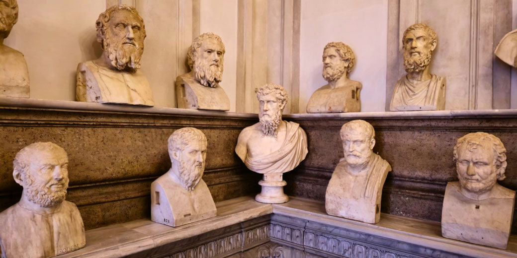 A collection of marble busts of ancient philosophers and statesmen displayed on tiered shelves, each with distinct facial features, beards, and expressions, arranged in a museum setting.