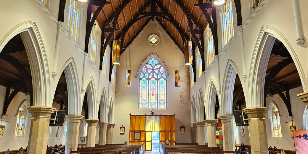 A church interior with arched columns, wooden ceiling, and a large stained glass window at the far end.