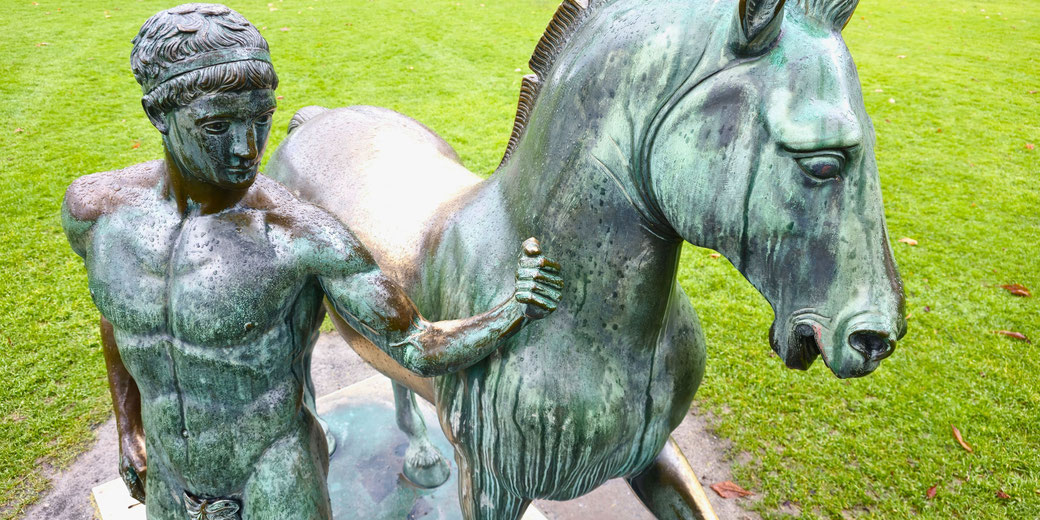 bronze statue of a muscular, nude man beside a horse, both detailed with weathered green patina. The statue is outdoors on a grassy area, with raindrops visible on the surface.