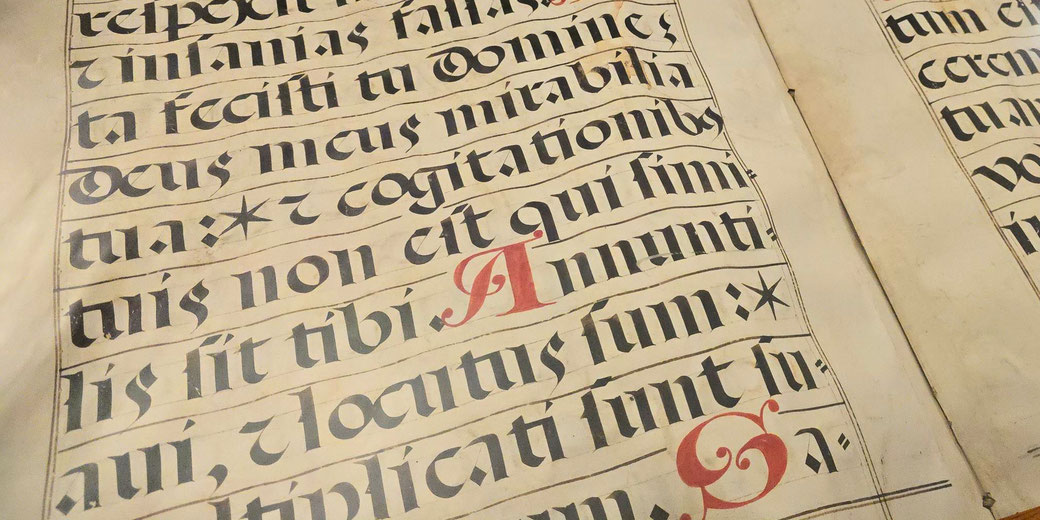 A medieval religious songbook with Latin script in black ink, featuring ornate red initials and musical notation on parchment, evoking the artistry of illuminated manuscripts.