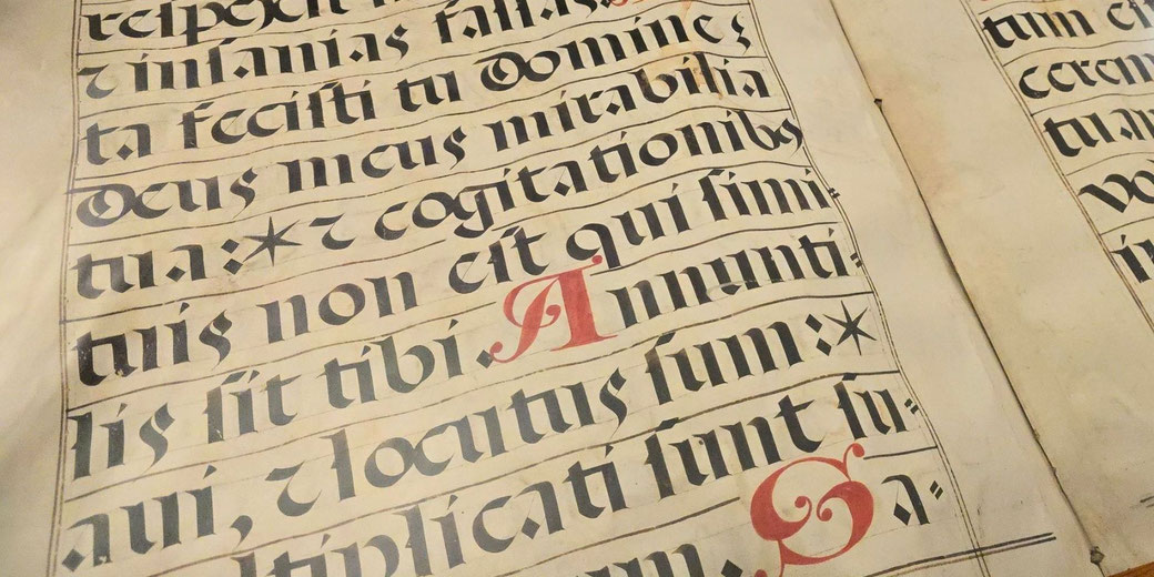 A medieval religious songbook with Latin script in black ink, featuring ornate red initials and musical notation on parchment, evoking the artistry of illuminated manuscripts.