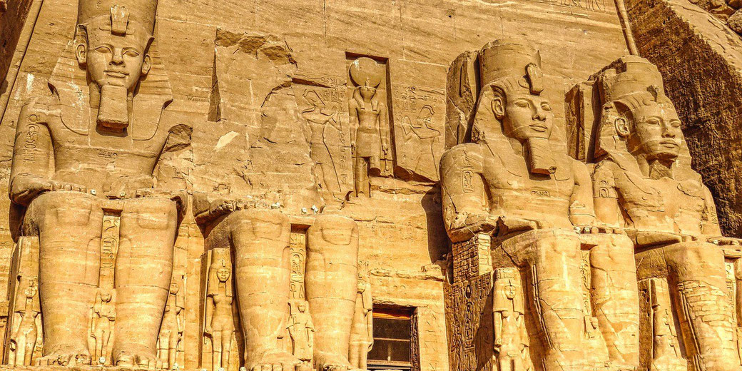Massive statues of Pharaoh Ramesses II carved into a sandstone cliff at Abu Simbel, with smaller figures and hieroglyphs surrounding them.
