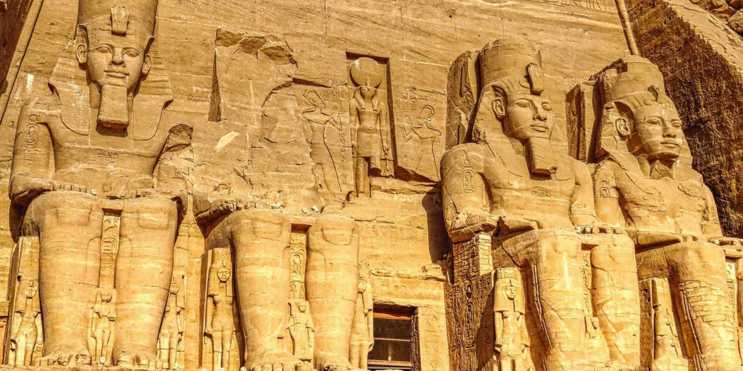 Massive statues of Pharaoh Ramesses II carved into a sandstone cliff at Abu Simbel, with smaller figures and hieroglyphs surrounding them.