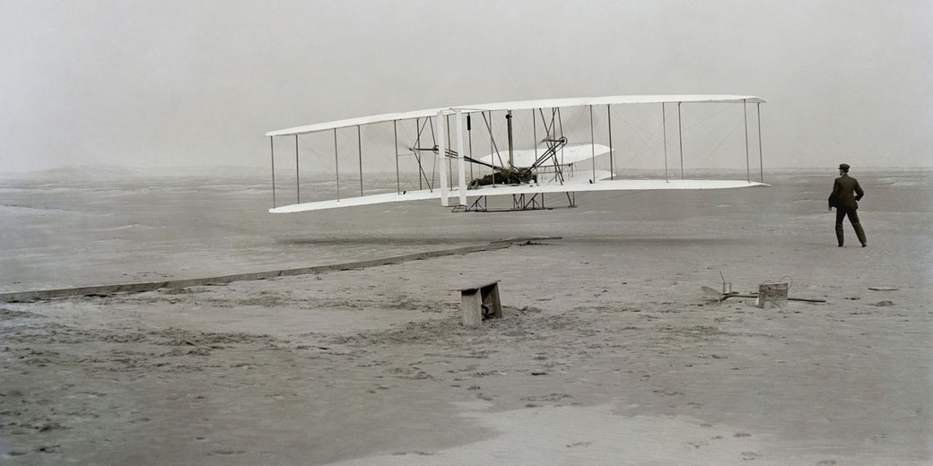 The Wright brothers' aircraft lifts off the ground in its first powered flight, with one brother piloting and the other observing from the sandy field.