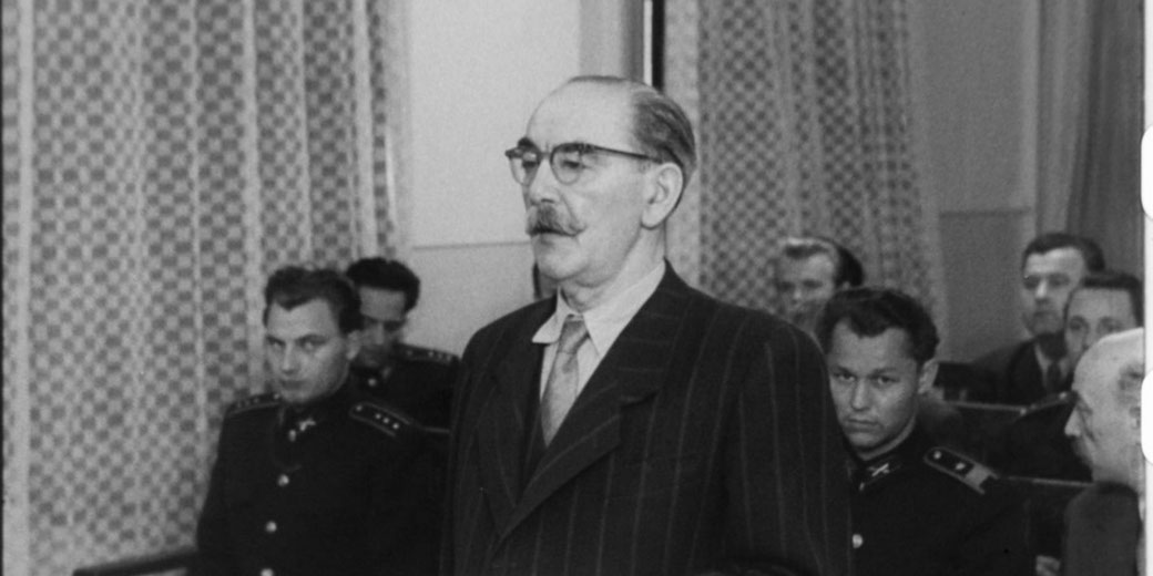 A black-and-white courtroom scene with a bespectacled man in a striped suit standing before seated officials and uniformed personnel, suggesting a formal legal proceeding.