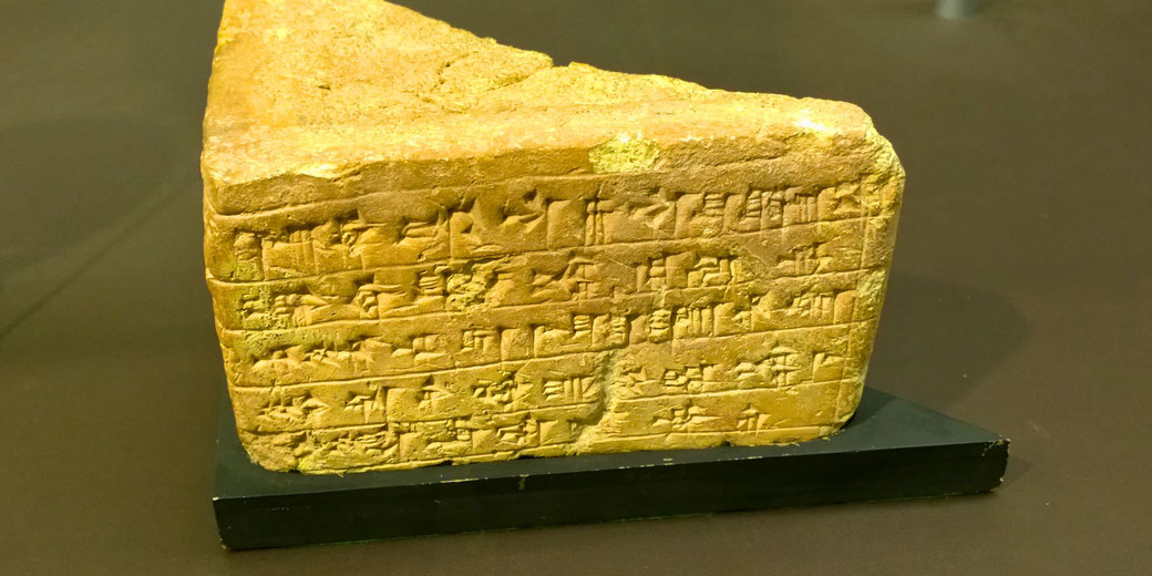 An ancient cuneiform-inscribed clay tablet with wedge-shaped writing. The artifact appears weathered and is displayed on a black stand, likely in a museum setting.