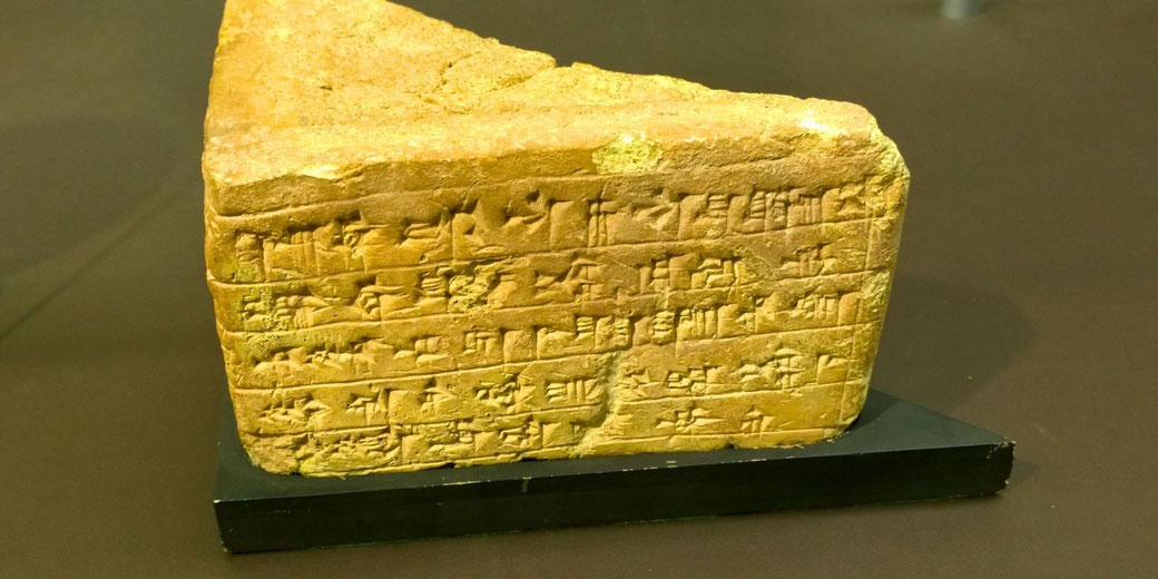 An ancient cuneiform-inscribed clay tablet with wedge-shaped writing. The artifact appears weathered and is displayed on a black stand, likely in a museum setting.