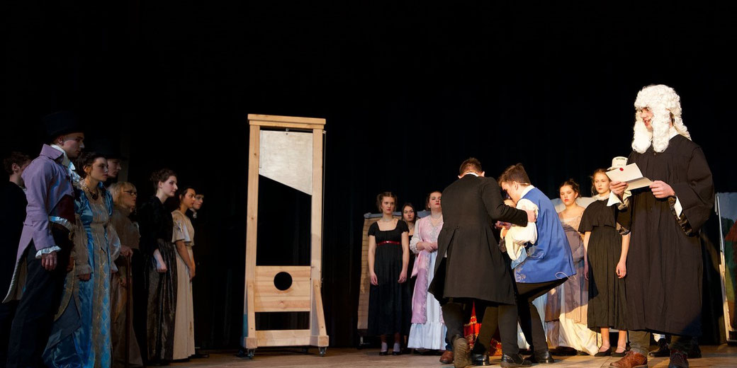 A group of actors in period costumes perform a dramatic guillotine scene on stage, with a judge reading and characters in tense poses.