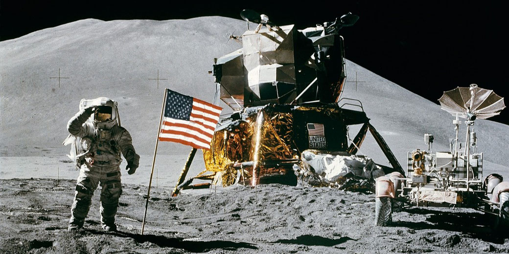 An astronaut salutes beside a U.S. flag, lunar module, and rover on the Moon’s surface under a dark sky.