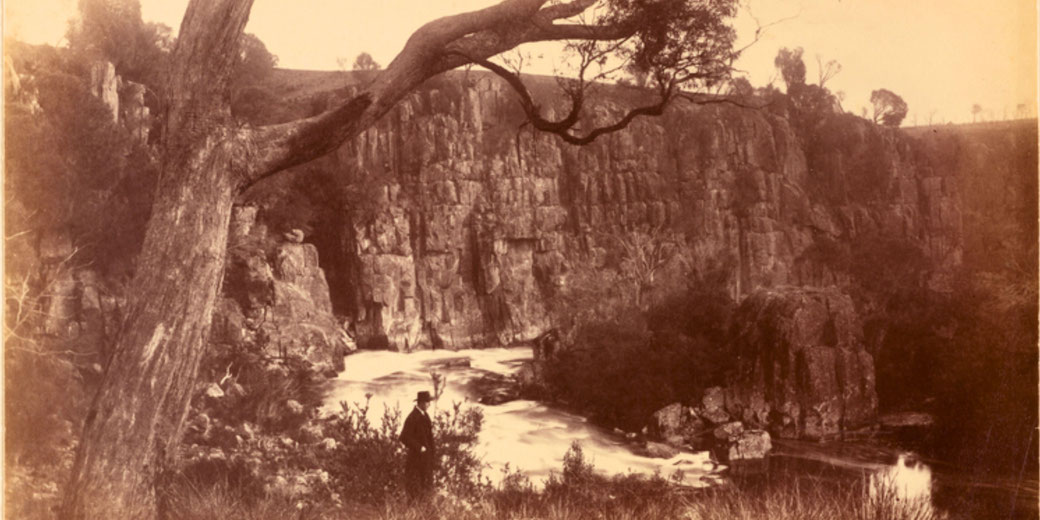 A sepia-toned photograph captures a rugged canyon with towering cliffs, a flowing river, and lush vegetation. A man in formal attire and a hat stands in the foreground.