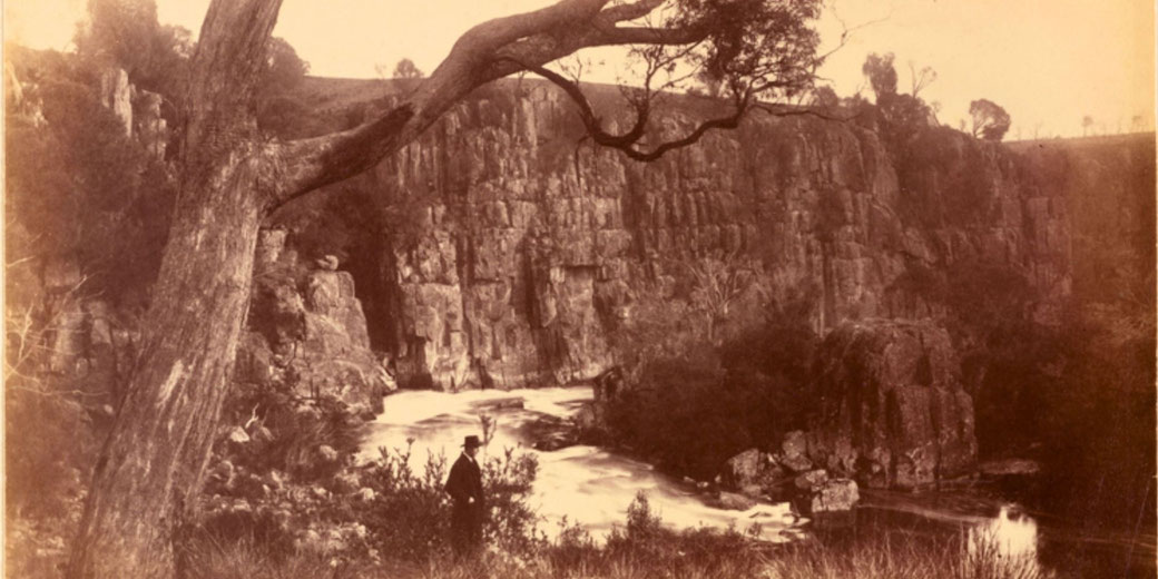 A sepia-toned photograph captures a rugged canyon with towering cliffs, a flowing river, and lush vegetation. A man in formal attire and a hat stands in the foreground.
