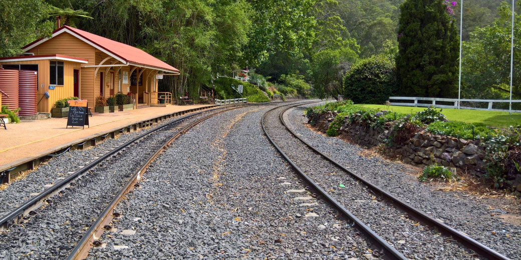 A quaint railway station with a wooden building, a red roof, and a platform beside curved train tracks.