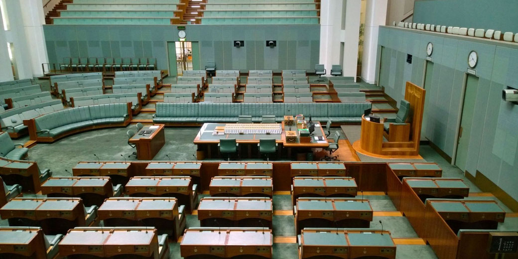 Interior of the Australian House of Representatives, featuring green benches, a central speaker's chair, desks with microphones, and a formal parliamentary layout.