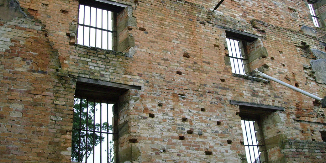 A crumbling brick building with barred windows, showing signs of decay and missing sections. The structure has exposed bricks, uneven surfaces, and a weathered appearance with trees visible through the windows.