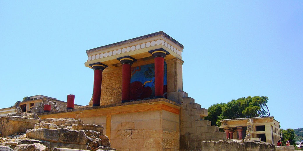 Reconstructed section of the Minoan Palace of Knossos with red columns, stone walls, and a colorful fresco under clear skies.