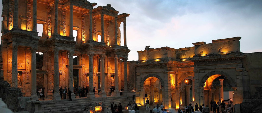 The illuminated ruins of the Library of Celsus and surrounding structures in Ephesus glow warmly at dusk with visitors below.