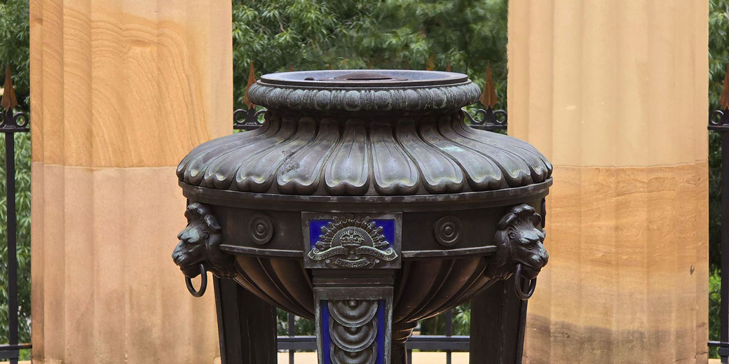 Bronze ceremonial urn with lion head handles and military insignia stands before stone columns and iron fencing.