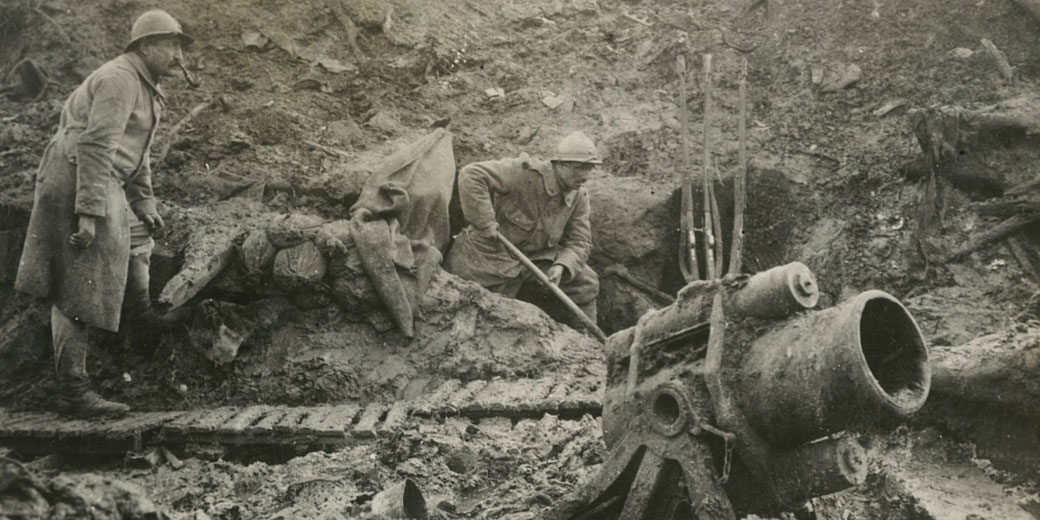 Two World War I soldiers work in a muddy trench near a large artillery piece; one smokes a pipe while the other uses a shovel.