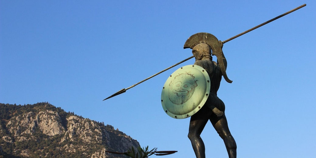 Bronze statue of an ancient Greek warrior holding a spear and round shield with a Medusa head, viewed from below against a clear sky and rocky hillside.