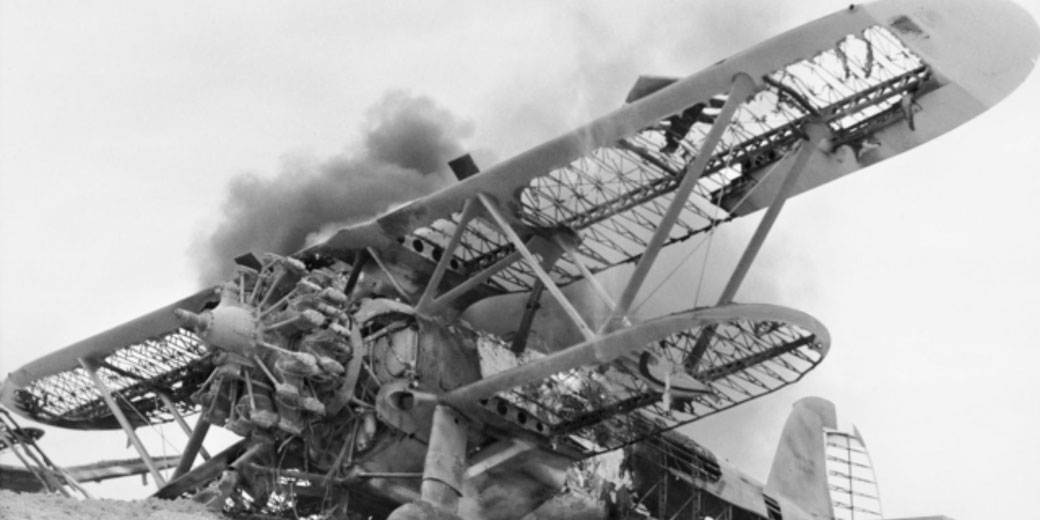 Damaged vintage biplane with broken wings and exposed engine emits thick smoke midair, captured in black and white with dramatic detail.
