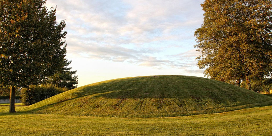A grassy burial mound surrounded by trees in a park-like setting.
