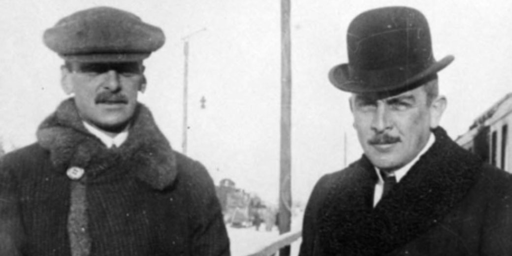 Two men in winter coats and hats stand outdoors near a train, with snow visible in the background.