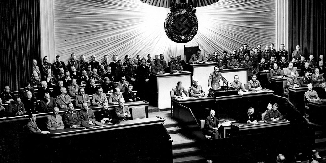 A large assembly of Nazi officials in uniform convenes in a grand hall under a swastika emblem, likely during a World War II-era governmental session.