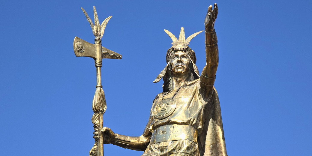 A golden statue of a Sapa Inca, wearing a headdress and armor, holding a ceremonial axe, and raising one hand, set against a clear blue sky.