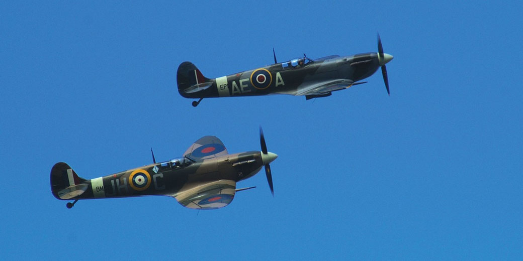 Two vintage British fighter planes fly in formation against a clear blue sky, showing roundels and wartime markings.