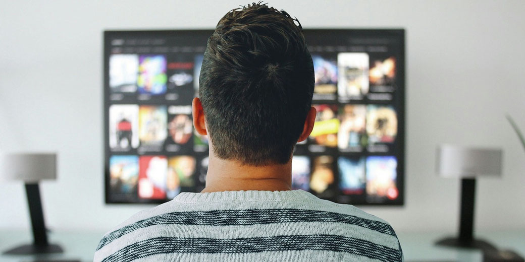 A person sitting in front of a TV screen browsing a streaming service, with various movie and show thumbnails displayed in a modern home setting.