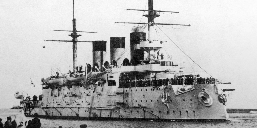 Black and white photo of an early armored warship at sea, packed with crew on deck, with tall masts, smokestacks, lifeboats, and heavy naval fittings.
