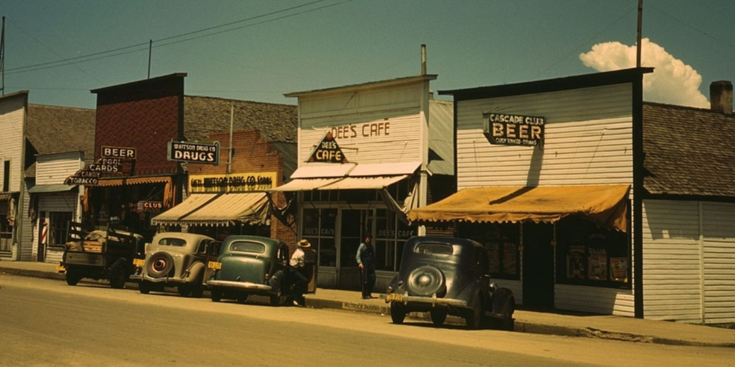 Small town street with 1930s cars parked outside cafés and shops under warm daylight skies.