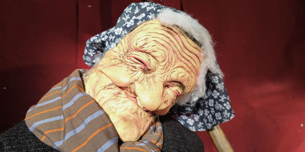 A close-up of a wrinkled, elderly-looking mask with exaggerated features, red eyes, and a mischievous grin.