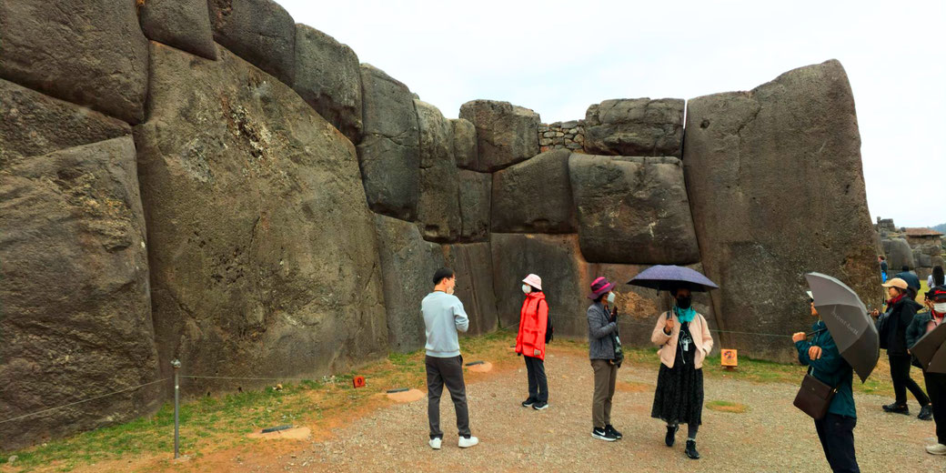 Tourists explore Sacsayhuamán, an ancient Inca site in Peru, featuring massive precisely-cut stone walls. Some visitors wear hats and masks, while others hold umbrellas on a cloudy day.