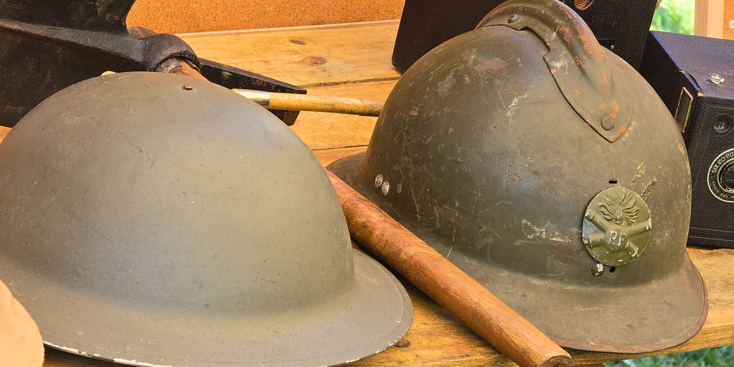 A British WWII Brodie helmet and a French WWII Adrian helmet on a wooden surface, along with vintage objects like wooden sticks and old cameras in the background.