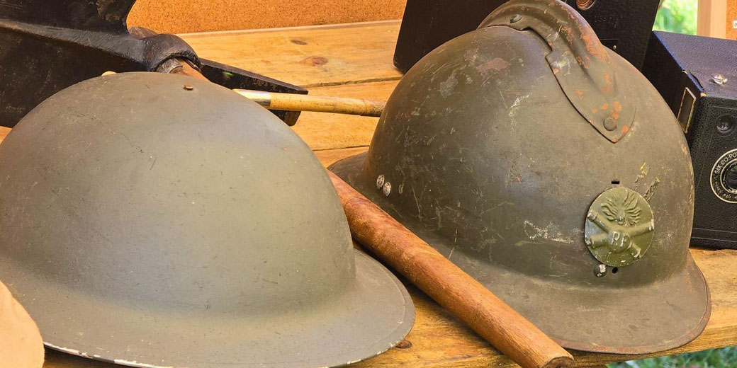 A British WWII Brodie helmet and a French WWII Adrian helmet on a wooden surface, along with vintage objects like wooden sticks and old cameras in the background.