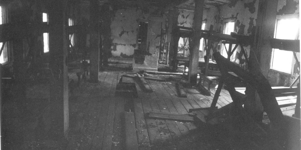 Dilapidated interior of a wooden building with broken floorboards, peeling walls, and scattered debris lit by daylight from the windows.