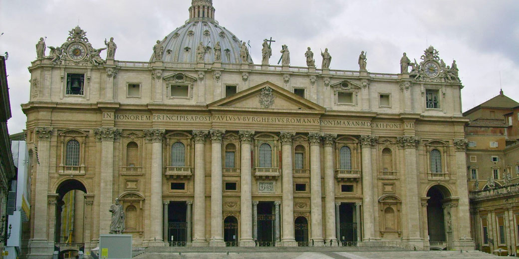 The grand façade of St. Peter's Basilica in Vatican City, featuring ornate columns, statues of saints, and a large central dome under a cloudy sky.