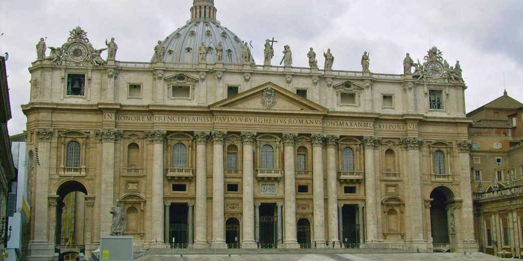 The grand façade of St. Peter's Basilica in Vatican City, featuring ornate columns, statues of saints, and a large central dome under a cloudy sky.