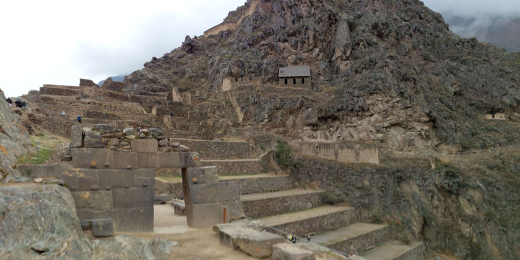 The ancient Inca ruins of Ollantaytambo in Peru, featuring stone terraces, a gateway, and a small stone structure on a rocky mountain under a cloudy sky.