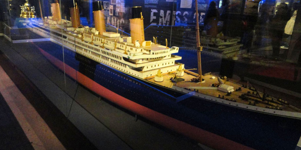 A detailed model of the RMS Titanic, displayed in a glass case at a museum, with surrounding exhibits visible in the background under dim lighting.