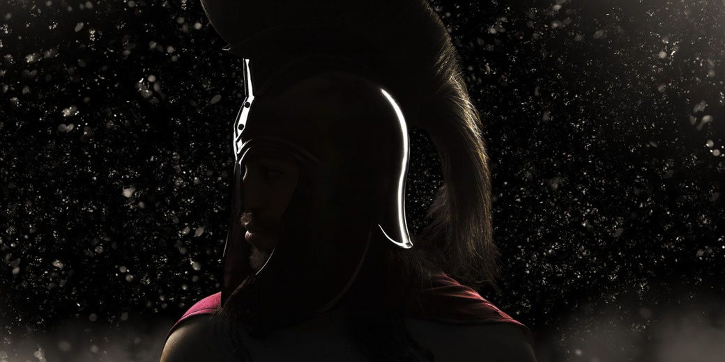 A Spartan warrior in profile, wearing a crested helmet and a red cape. Dramatic lighting highlights the helmet's shine, with dust or embers in the dark background enhancing the atmosphere.