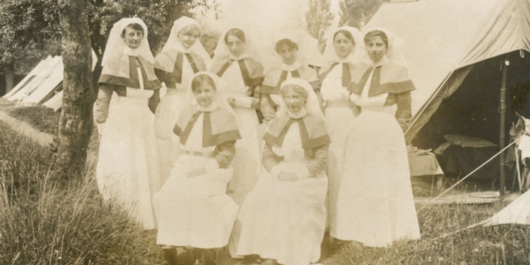 Eight nurses in early 20th-century uniforms pose in front of canvas tents, likely in a wartime medical camp setting.