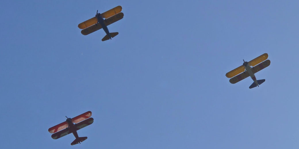 Three vintage biplanes with colorful wings fly in a triangular formation against a clear blue sky.