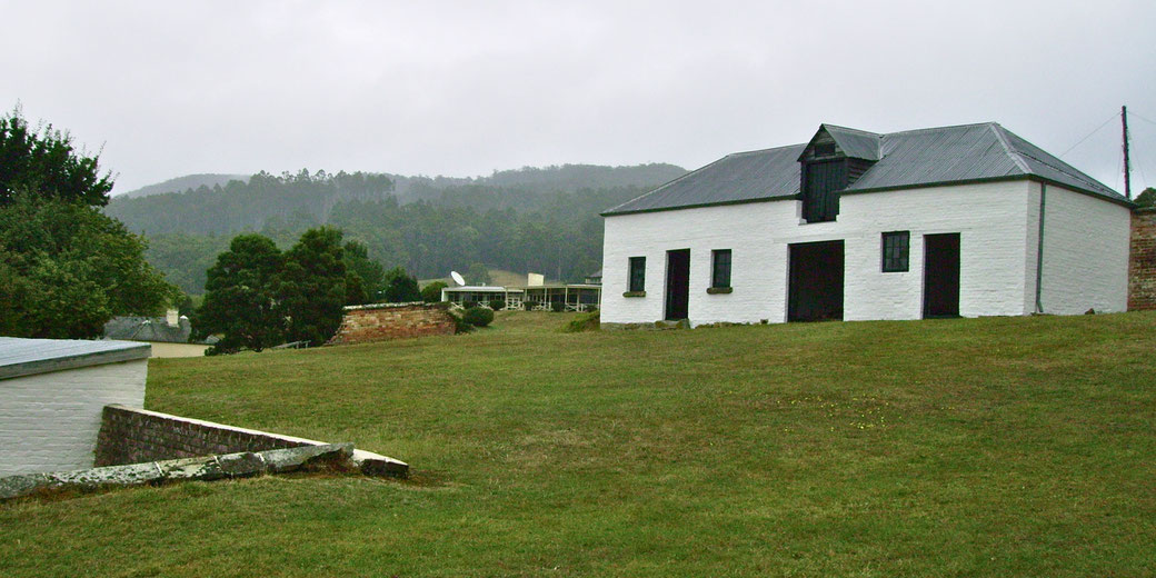 A white brick building with a metal roof sits on a grassy hill, surrounded by trees and distant mountains under an overcast sky.