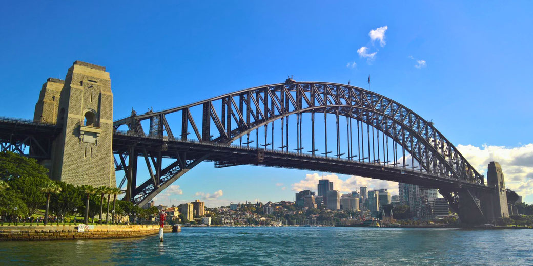 Iconic steel arch bridge spanning across water with stone pylons, urban skyline, and blue sky in the background.