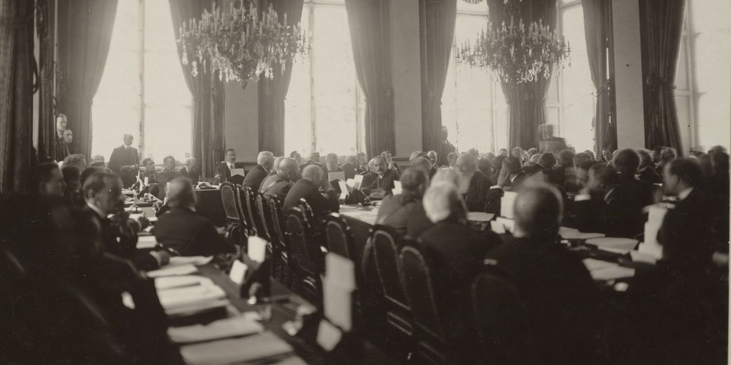 Formal meeting of suited men seated at long tables in a grand hall with tall windows, heavy curtains, and large chandeliers, suggesting an early 20th century conference.