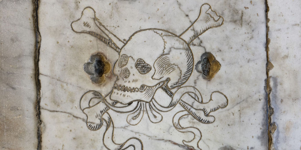 Engraved skull and crossbones with flowing ribbons on aged marble, flanked by two metal bolts, symbolizing death or danger.